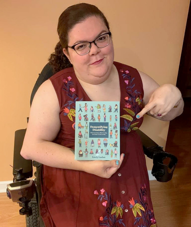 Emily Ladau, a white woman wearing glasses and a dress embroidered with flowers, holds and points to a copy of Demystifying Disability while sitting in her motorized wheelchair