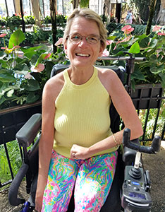Becky, smiling, in a botanical conservatory wearing summer clothes and using her power chair