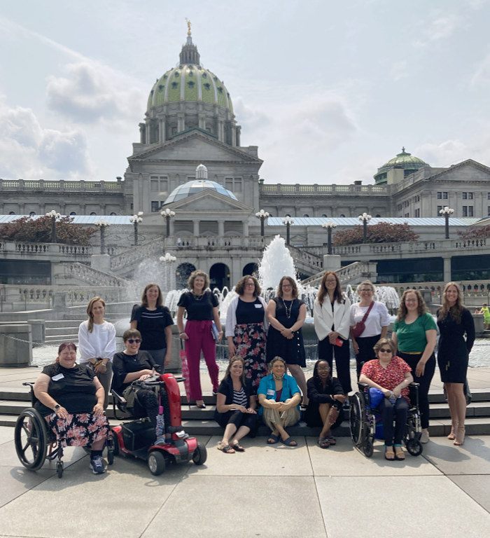 C2P2 cohort of 15 women outside of the Capitol stand, sit, or use wheelchairs or scooters near a fountain in front of Capitol