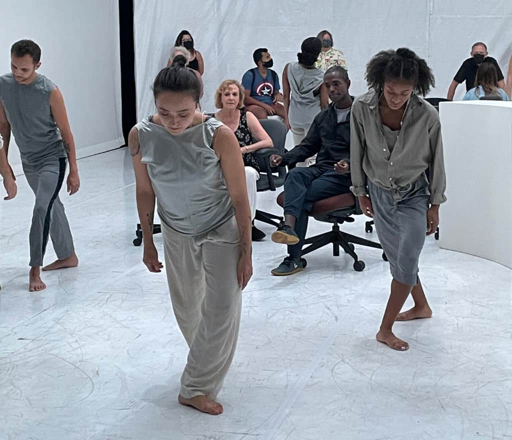 3 dancers in gray garments in a white space lean forward in a triangular formation, watched by audience within the space