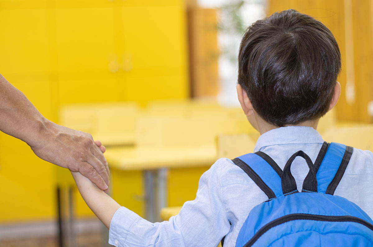 Young child approaches classroom wearing backpack and holding an adult's hand.