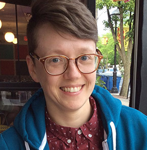 Dynah, smiling, stands outdoors near a building wearing glasses and casual clothes