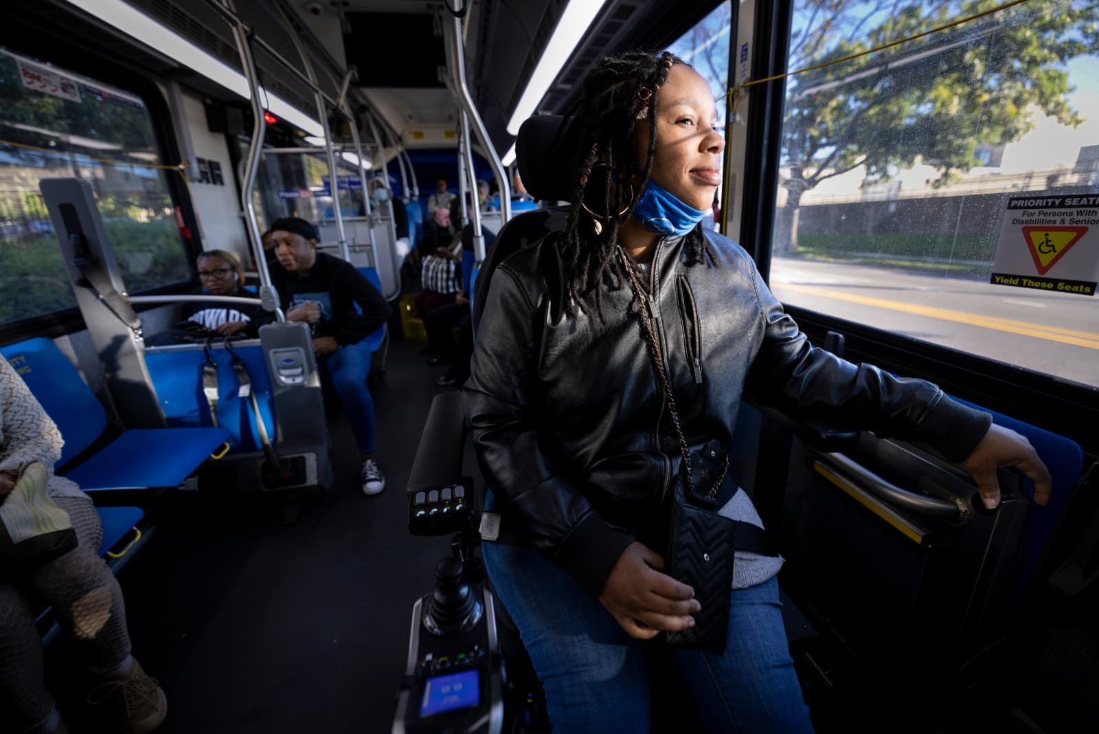 Domonique gazes out the window of a public transit bus, smiling slightly, while perched in her power wheelchair in the accessible area at the front of the bus.