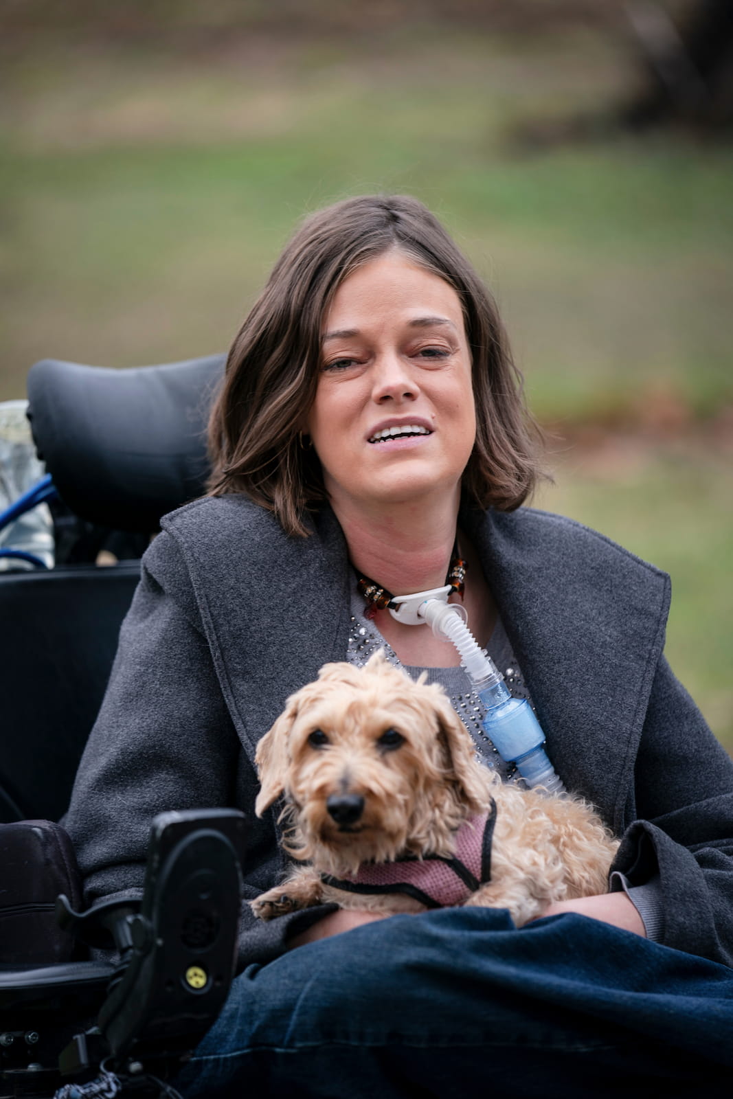 Josie, a white woman with medium length brown hair holds her small dog in her lap outside in her wheelchair. She uses a breathing tube