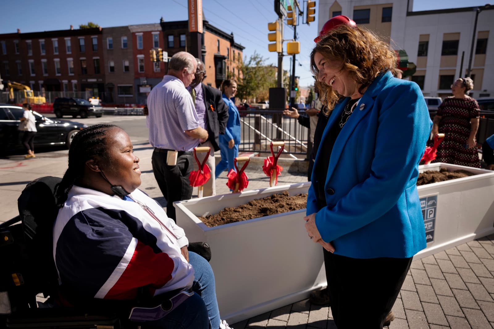 On a city sidewalk next to a planter filled with fresh soil, a Black woman using a wheelchair and a white woman wearing a suit chat, among other small groups