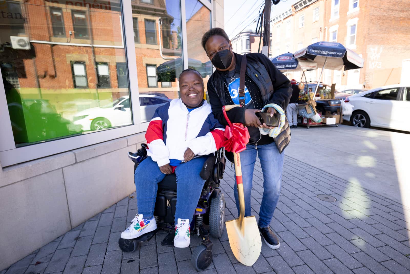 LaToya, on the sidewalk in Philadelphia, poses with a woman, a gold shovel with a red bow balanced between them