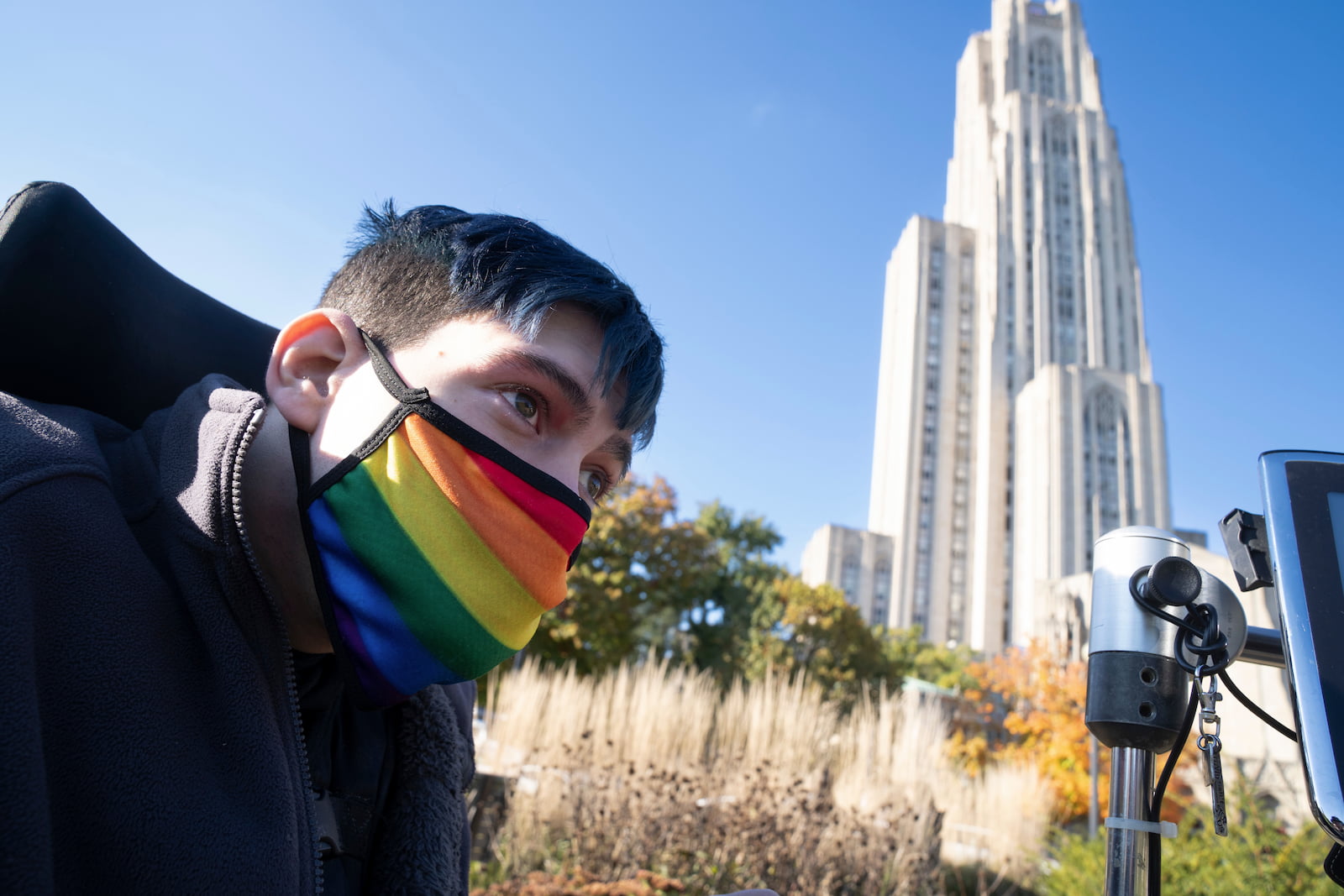 Close-up of Mark outdoors wearing a rainbow face mask with his AAC device in view and a tall building beyond