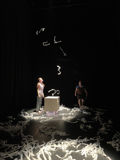 Paper strips float above 3-foot tall light-filled cube centered in a dark room with more strips scattered on the floor, 2 women watching the paper above them