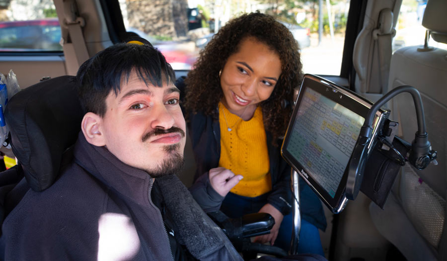 Young disabled man with a satisfied expression inside a vehicle uses a tablet for augmentative communication and is accompanied by his assistant, a Black woman