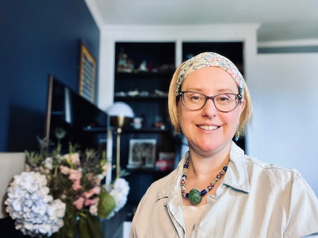 Sarah Holland, smiling, stands near flower arrangement and wears glasses, chunky jewelry, and a wide headband holding back short hair.