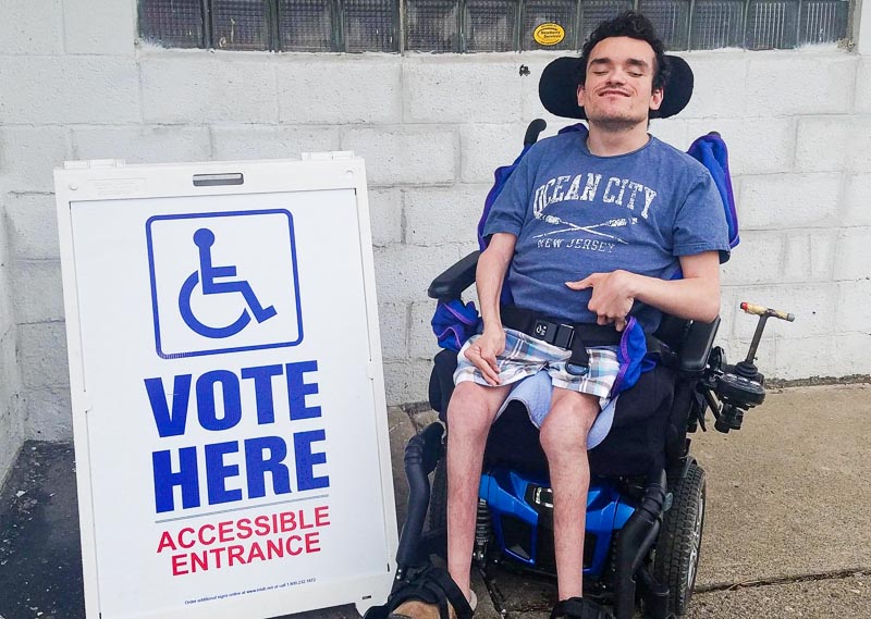 Robert using wheelchair next to Vote Here, Accessible Location sign at a polling place
