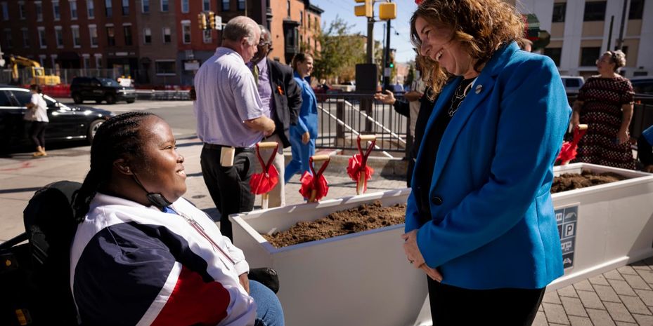 On a city sidewalk next to a planter filled with fresh soil, a Black woman using a wheelchair and a white woman wearing a suit chat, among other small groups