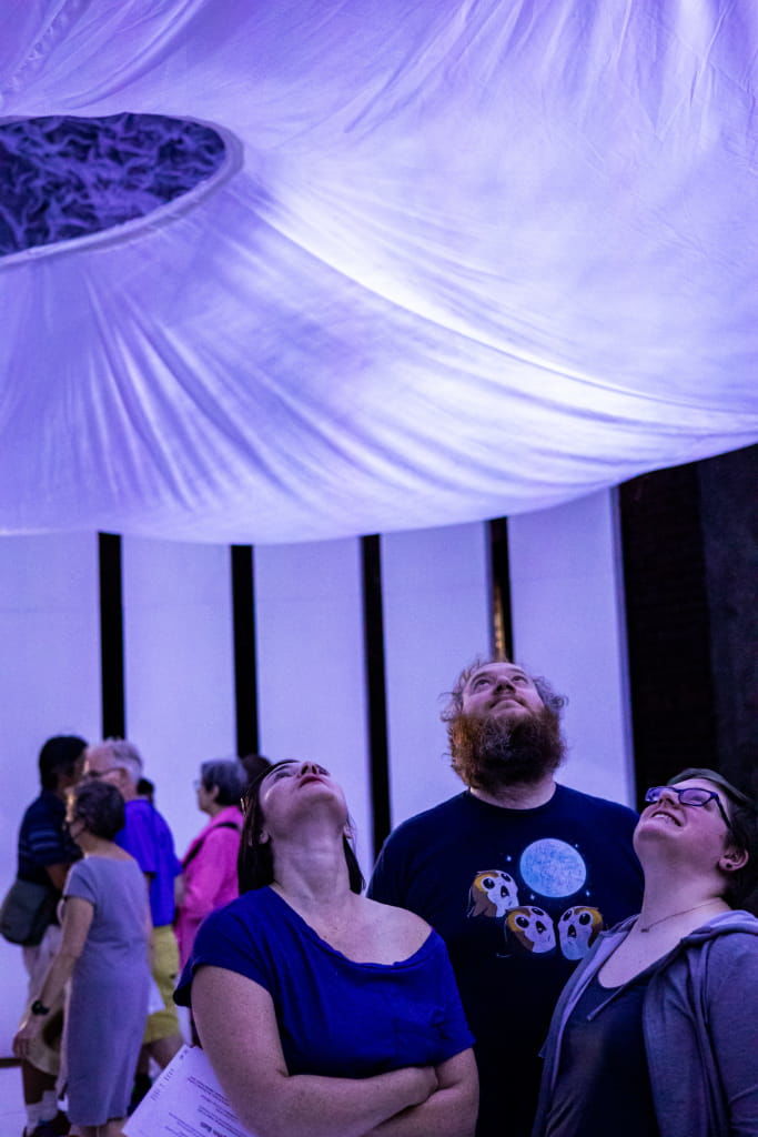 3 audience members gaze up at white fabric ceiling that's lit from within