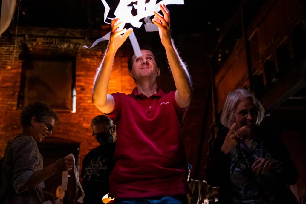 a man in a dimly lit space, among others, holds up strips of spot-lit white paper 