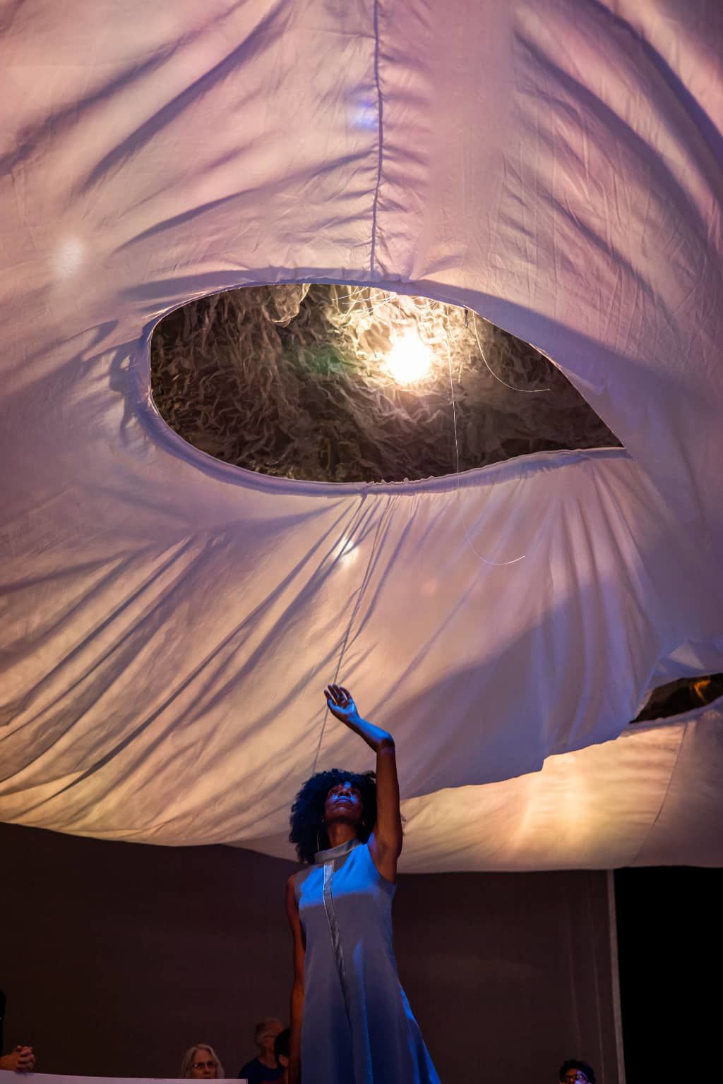 dancer partially lit with blue light reaches upward toward a round opening in backlit white fabric ceiling