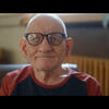 An elder man wearing glasses looks straight ahead while sitting indoors, backlit by window light