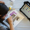 A student leans over their painting on paper in progress, with their tablet standing nearby on the table displaying an AAC app with buttons and words for colors