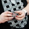 A person with red, white, black, and silver-painted nails holds a white, red, and black pin with the text C2P2