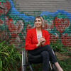 Woman with short blond hair and red sweater using wheelchair in outdoor garden space bordered by mural of strawberries painted on a brick wall