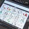 An AAC device with grid of images and corresponding text. Food is represented by a loaf of bread and an apple. I need help is pictured by a person reaching out to someone on the ground.