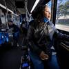 Domonique gazes out the window of a public transit bus, smiling slightly, while perched in her power wheelchair in the accessible area at the front of the bus.