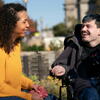 Young man using power wheelchair outdoors in community with assistant