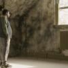 Jonathan stands in a room at Pennhurst. He looks out a window at trees. The walls of the room bloom with mold. The window casts light on Jonathan, whose shadow falls behind him, toward an open door.