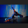Two performers on a stage dimly lit in blue, connect via one's hands and the other's wheels, as the wheelchair user lays back with arms stretched and hands resting on the stage. The other balances with straight arms, in a flying position along the tipped-up wheelchair