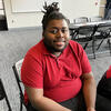Zhyir, a youthful Black man with tied-back locs and wearing a red polo shirt, sits in a conference room, smiling at the camera