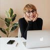Smiling adult with short hair and glasses leans elbows on table in front of their open laptop