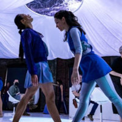 Two dancers face each other in a dimly lit space below a billowing white ceiling, one leaning forward, the other backward