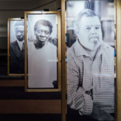 View of exhibit shows several black and white photo portraits printed on vertical banners. The expressions are serious.