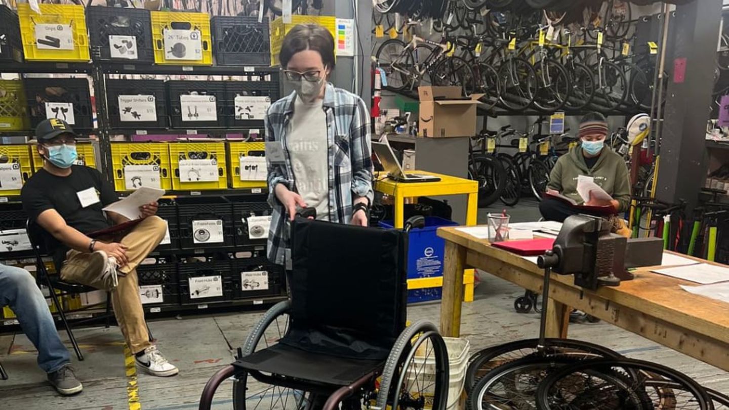 Caitlin stands in bike shop with wheelchair, surrounded by shop equipment and several people