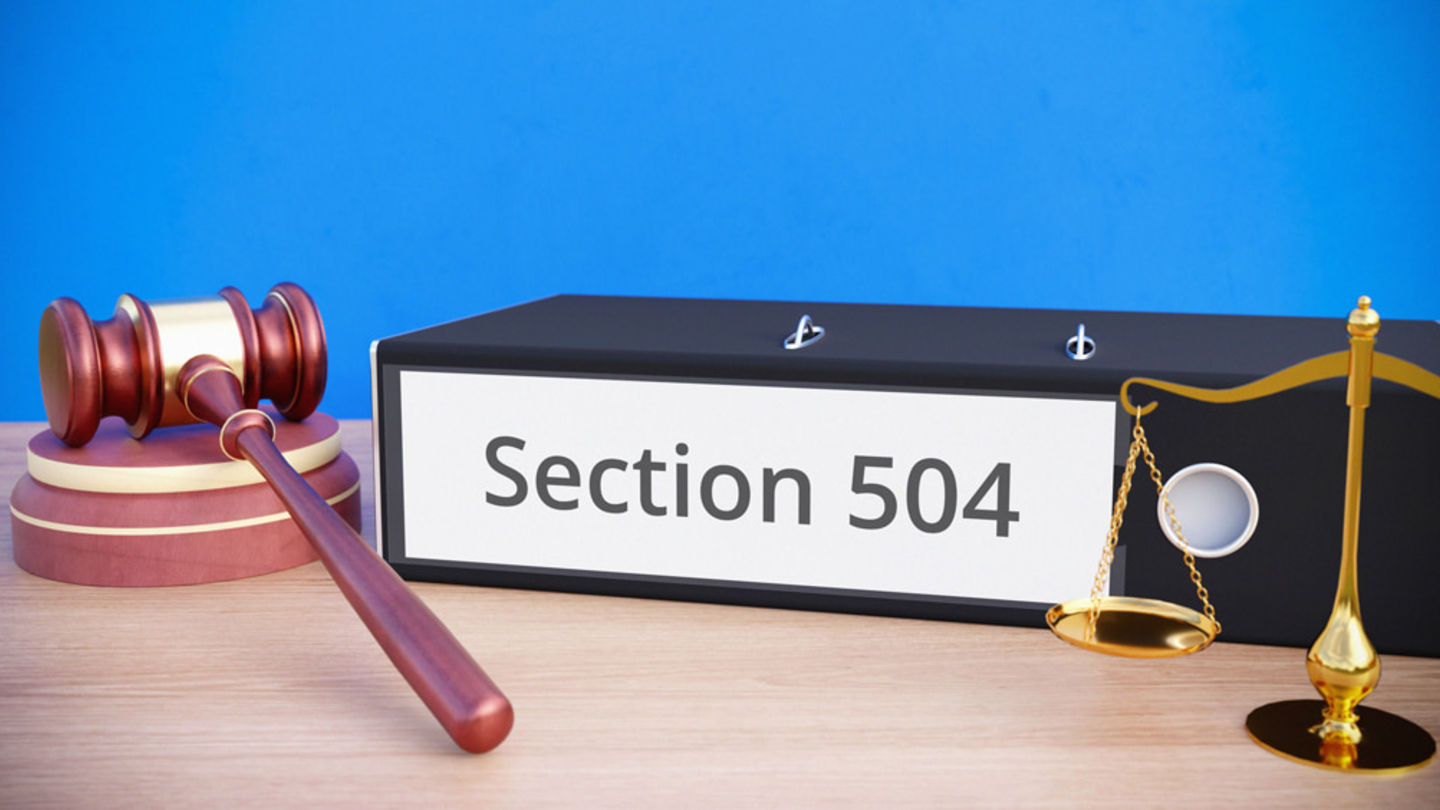 gavel rests on table beside a binder with Section 504 printed on its spine and a nearby scale of justice