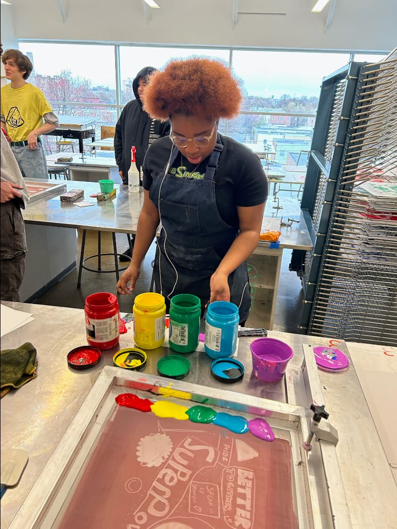 Person wearing apron has ink jars open and brightly colored ink added to a silkscreen, ready to squeegee